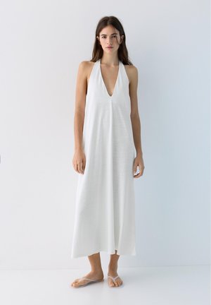 Jeune femme debout dans une robe blanche sans manches à col halter et des sandales transparentes à enfiler, sur un fond uni.