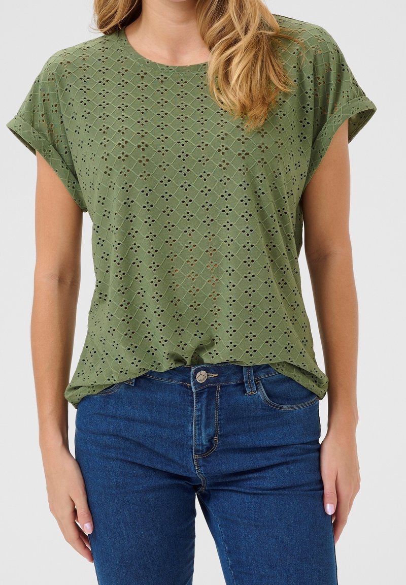 Femme portant un haut vert à manches courtes avec des œillets et des revers roulés, ainsi qu'un jean bleu taille haute, sur un fond uni.