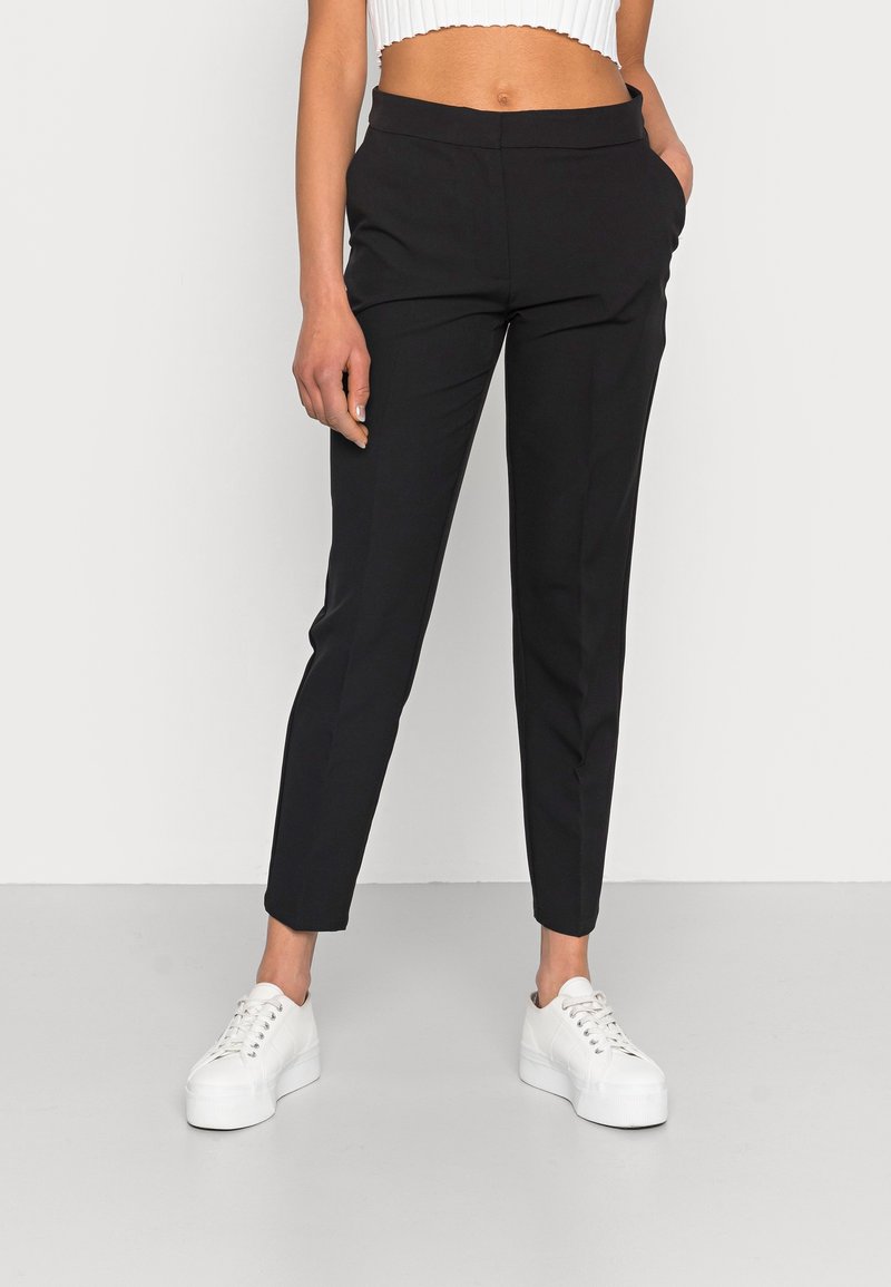 Pantalons fuselés noirs avec poches latérales, présentant une texture de tissu lisse et un design minimaliste. Portés avec des baskets à plateforme blanches.