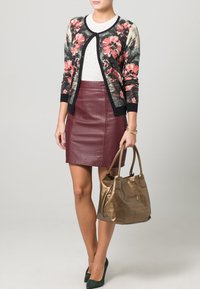 Donna che indossa un cardigan floreale, una maglietta bianca, una gonna di pelle bordeaux, tacchi verdi e tiene una borsa metallica bronzo.