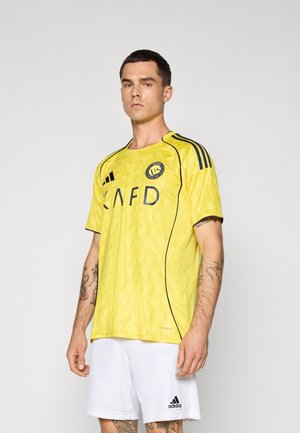 Geel sportshirt met golvend textuur, zwarte accenten en 'CAFD'-logo, gecombineerd met witte shorts en zwart Adidas-logo op de shorts.