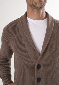 Pruunist lõngast kootud cardigan uhke kraega, millel on tekstuuritooted ribid, kaks musta nööpi ja mis on kihiliselt valge särgi kohal.