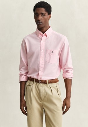 Mann trägt ein hellrosa Hemd mit Brusttasche, beige plissierte Hose und braunen Gürtel, steht vor einfarbigem Hintergrund.