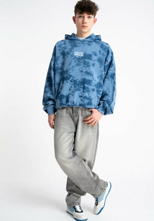 Nastolatek w niebieskiej bluzie z kapturkiem tie-dye z napisem "Endless Summer", szarych luźnych dżinsach oraz białych tenisówkach z niebieskimi akcentami, stojący ze skrzyżowanymi nogami.