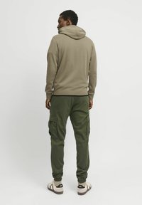 Person står med ryggen til iført en beige hættetrøje, grønne cargobukser og hvide sneakers mod en ensartet lys baggrund.