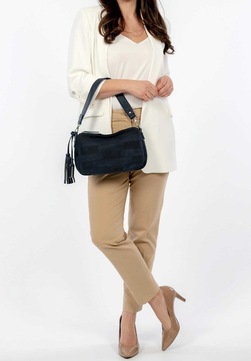 Czarna tekstylna torba na ramię o zaokrąglonym kształcie, z zamkiem błyskawicznym i detalami frędzli. Noszona z beżowymi spodniami i białą marynarką.