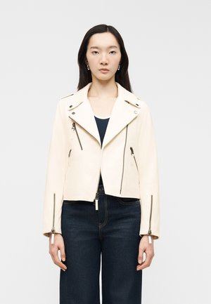 Woman with long dark hair wearing a cream biker jacket over a dark top and dark jeans, standing against a plain light background.