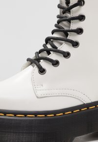 Botte plateforme en cuir blanc avec semelle épaisse noire, surpiqûres jaunes et lacets noirs passant dans des œillets métalliques.