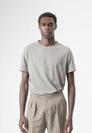 Hombre de pie mirando al frente, vestido con una camiseta gris claro y pantalones beige plisados sobre un fondo blanco liso.