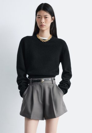 Schwarzer Strickpullover mit hohem Kragen, kombiniert mit hoch sitzenden, grauen, maßgeschneiderten Shorts. Ergänzt durch eine auffällige goldene Kettenhalskette.