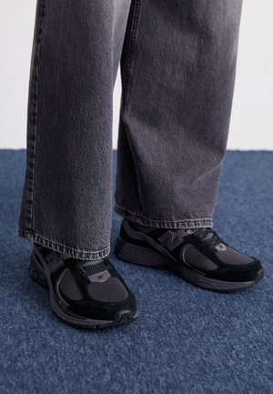 Chaussures de sport noires et grises avec des matériaux en maille et en daim, présentant un logo distinctif et des semelles rembourrées, associées à un pantalon gris.