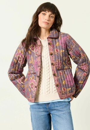 Kvinde iført en lilla blomstret quiltet jakke over en cremefarvet kabelstrikket sweater og blå jeans, stående med hænderne på hofterne mod en ensfarvet baggrund.