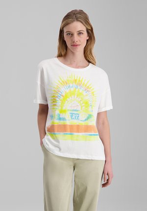 Frau mit langen blonden Haaren trägt ein weißes T-Shirt mit buntem Sonnenstrahl und dem Text "LIBERTY", kombiniert mit hellbeigen Hosen vor schlichtem grauem Hintergrund.