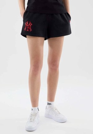 Person trägt schwarze Shorts mit rotem Logo auf der linken Seite und weiße Sneakers mit weißen Socken und steht vor einem einfarbigen Hintergrund.