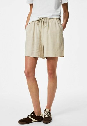 Személy bézs színű húzózsinóros rövidnadrágot, fehér pólót és barna tornacipőt visel, kezét zsebében tartva áll fehér háttér előtt.