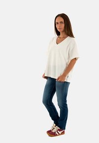 Vit V-ringad topp med korta ärmar, gjord av lätt tyg, kombinerad med tajta blå jeans och vinröda sneakers med vita detaljer.