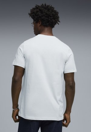 Uomo con capelli ricci e occhiali, indossa una t-shirt bianca a maniche corte e pantaloni scuri, in piedi di spalle su uno sfondo grigio.