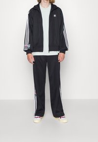 Czarny sportowy dres z białymi paskami, bluza na zamek, spodnie z elastycznym pasem, ozdobione wielokolorowymi naszywkami z logo oraz jasno niebieska koszula pod spodem.