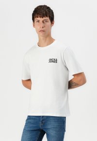 Hvit bomull t-skjorte med korte ermer, med en svart logo "JACK & JONES" på venstre bryst. Båret med blå jeans.