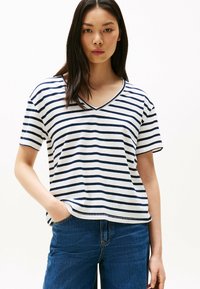 Joven mujer con cabello largo y oscuro, vistiendo una camiseta de rayas blancas y marinas en V y jeans azules, de pie con una mano en el bolsillo.