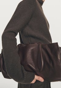Brązowa skórzana torba o gładkim wykończeniu, z wydłużonymi uchwytami i usztywnionym kształtem, trzymana pod oliwkowozielonym dzianym swetrem.