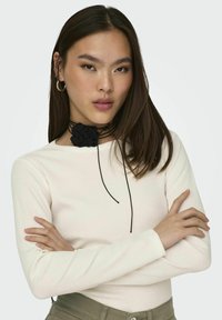 Zwarte bloemen choker op een model met lang bruin haar, die een wit lange mouwen top draagt. De choker heeft meerdere zwarte strengen en een textuurbloem.