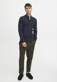 Cămașă albastră cu nasturi, cu mâneci lungi, asortată cu pantaloni chino verde măsliniu și pantofi negri, având o textură netedă și un design ajustat.