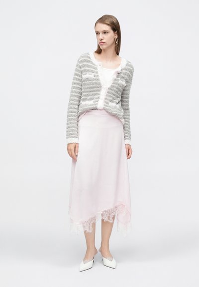 Γυναίκα που φοράει γκρι και λευκή ριγέ ζεστή ζακέτα πάνω από λευκό τοπ, φούστα απαλό ροζ με δαντελωτό τελείωμα και λευκά μυτερά mule παπούτσια.