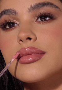 Lśniący, nude różowy błyszczyk do ust nałożony aplikatorem, podkreślający pełne usta i subtelny blask. Brązowe oczy i wyraźne brwi w zbliżeniu.