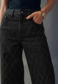 Černé džíny z denimu s diamantovým vzorem vytvořeným malými vsazenými cvočky. Mají vysoký pas a volný střih s předními kapsami.