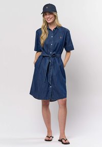 Džínové košilové šaty v tmavě modré barvě s krátkými rukávy, knoflíky vpředu a páskem na zavázání v odpovídající barvě. Doplňují je tmavě modrá džínová čepice.
