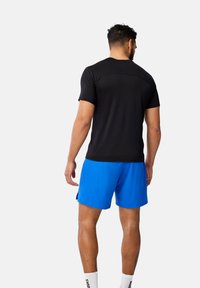 Svart kortärmad atletisk tröja med en slät textur, i kombination med blå atletiska shorts, bärs av en person som ses bakifrån.