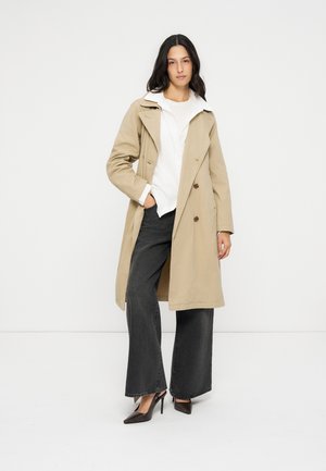 Mujer con un abrigo beige tipo trench sobre una camiseta blanca, vaqueros negros de pierna ancha y zapatos de tacón negros, de pie sobre un fondo blanco.
