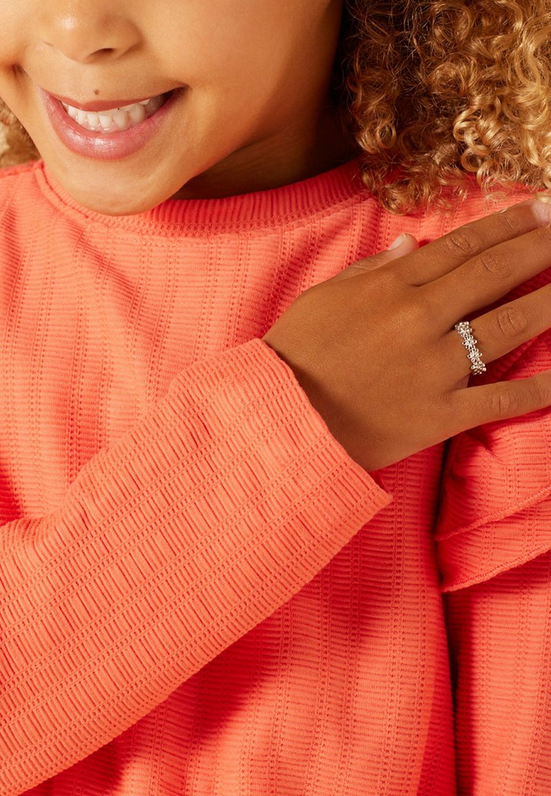Heldere koraalkleurige lange mouw top met een textuurpatroon. Zilveren ring met bloemontwerp gedragen op een hand die op de mouw rust.
