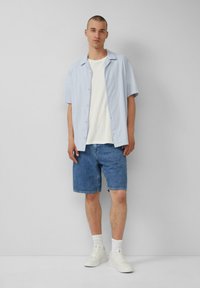 Lichtblauwe t-shirt met korte mouwen over een wit T-shirt, gecombineerd met denim shorts en witte sneakers. Eenvoudig ontwerp met knoopdetails.