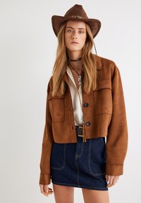 Veste cropped en suédine marron avec deux poches avant, boutons noirs et une ceinture. Portée sur une chemise claire et assortie à une mini-jupe en denim.