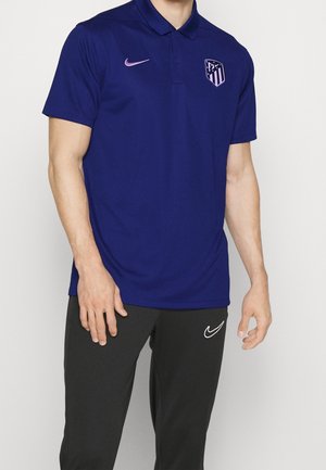 Man wearing a blue polo shirt with Atlético Madrid logo and black Nike pants with white swoosh on the left thigh.
