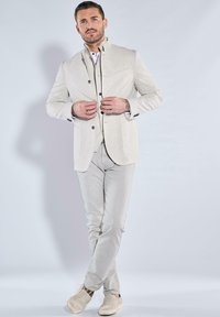 Helles, beiges Leinenblazer mit dunklen Knöpfen, kombiniert mit einem weißen Kragenhemd und hellgrauen Hosen, ergänzt durch beige Slipper.