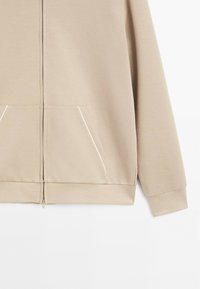 Haut à capuche beige zippé en tissu doux, avec deux poches avant bordées de blanc, des poignets côtelés et un ourlet droit.