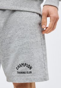 Άτομο που φορά γκρι σορτς με την επιγραφή «CHAMPION TRAINING CLUB» σε μαύρο στο κάτω μέρος του μηρού, το χέρι ακουμπά κοντά στην τσέπη.