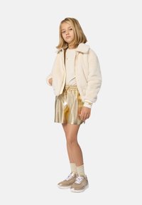 Fluffy crème bomberjack, gouden metallic shorts, lichtbeige top, gouden sneakers en beige sokken. Glad textuur en ontspannen pasvorm.