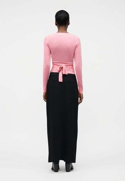 Femme à la peau foncée et aux cheveux courts portant un haut rose à manches longues noué dans un nœud dans le dos et une jupe noire longue jusqu'aux chevilles avec des chaussures noires.