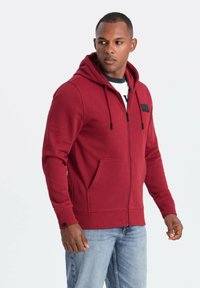 Sudadera con cremallera roja, bolsillos tipo canguro, capucha ajustable con cordón, puños acanalados y un parche de logo negro en el pecho.