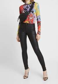 Persoon in zwarte hoog taille nepleer broek, kleurrijke lange mouwen met patroon top, zwarte schoudertas en zwarte bandjes met hoge hakken sandaaltjes.