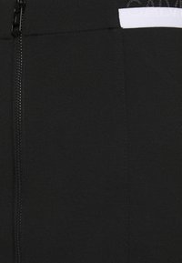 Svart tyg med en vertikal dragkedja på vänster sida och en liten delvis vit etikett med svart text i det övre högra hörnet.