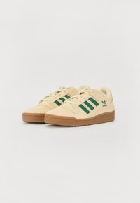 Beige sneakers laget av semsket skinn og gummi. Har tre grønne striper, polstret krage og en gummisåle; diskret merkevare på siden.