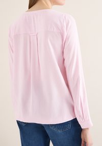 Femme portant un chemisier à manches longues blanc à rayures rose clair et un jean bleu, vue de dos sur un fond uni.