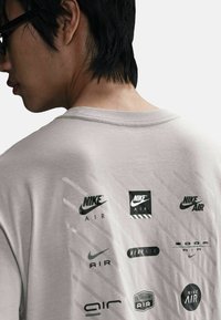 Camiseta gris claro con múltiples logotipos de Nike impresos en negro en la parte trasera, con cuello redondo y mangas cortas.