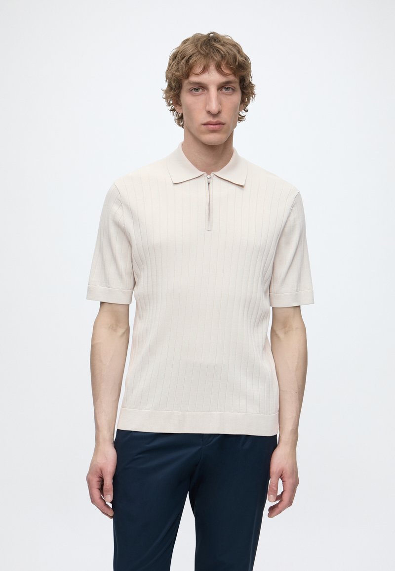 Jeune homme aux cheveux bouclés portant un polo en maille beige à manches courtes avec une fermeture éclair à l'avant et un pantalon bleu marine foncé, debout devant un fond blanc.