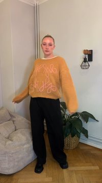Overdimensjonert oransje genser med rosa kursiv broderi, kombinert med svarte vide bukser og svarte sko, stående ved siden av en beige sofa.